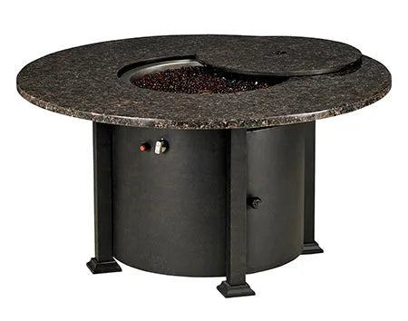 A round outdoor fire table with a granite top and a black metal base. The table has a circular burner and a control panel with a knob and a switch. A granite lid covers the burner, and a door on the side of the base reveals a storage compartment.