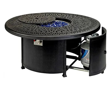  A round outdoor fire table with a woven base and a travertine stone tabletop. The table has a circular burner and blue flames are visible. There is a propane tank storage compartment at the base, and the table has four small wheels. The image shows a side view of the table with the storage compartment door open, revealing the propane tank inside.