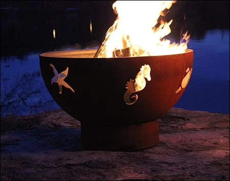 A round, rusted metal fire pit with flames burning inside. The fire pit has seahorse, crab, and fish designs cut into the metal. It is placed on a rock near a body of water at night.