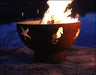 A round, rusted metal fire pit with flames burning inside. The fire pit has seahorse, crab, and fish designs cut into the metal. It is placed on a rock near a body of water at night.