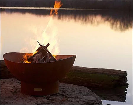 A round, rusted metal fire pit with flames burning inside. The fire pit is placed on a rock near a body of water at sunset. The sky is orange and the water is reflecting the flames.