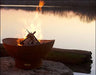 A round, rusted metal fire pit with flames burning inside. The fire pit is placed on a rock near a body of water at sunset. The sky is orange and the water is reflecting the flames.