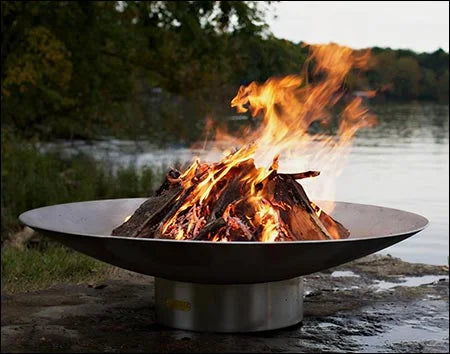 A large, round stainless steel wood burning fire pit with a roaring fire in the center. The fire pit is placed on a rocky shoreline near a lake with lush greenery in the background.