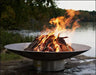 A large, round stainless steel wood burning fire pit with a roaring fire in the center. The fire pit is placed on a rocky shoreline near a lake with lush greenery in the background.