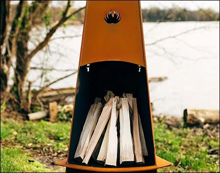 A tall, conical fire pit with firewood inside. The fire pit is surrounded by green grass and is near a body of water. There is a deer logo on the side of the fire pit.