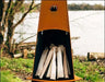 A tall, conical fire pit with firewood inside. The fire pit is surrounded by green grass and is near a body of water. There is a deer logo on the side of the fire pit.