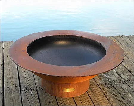 Wood burning fire pit on a wooden deck by a lake. The fire pit is round and made of rusted metal. There is a small light on the bottom of the fire pit.