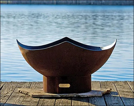 A large, round fire pit made of rusted metal is sitting on a wooden dock. The fire pit has a unique, manta ray-shaped design. A body of water is visible in the background.