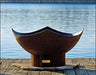 A large, round fire pit made of rusted metal is sitting on a wooden dock. The fire pit has a unique, manta ray-shaped design. A body of water is visible in the background.