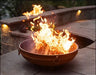 A round, rusted metal fire pit with a large, roaring fire burning inside. The fire pit has two handles on the side.