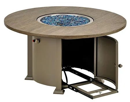 A round gas fire pit table with a wooden top and a bronze-colored base. The fire pit has blue flames and glass fire beads. A door on the side of the base is open, revealing a storage compartment.