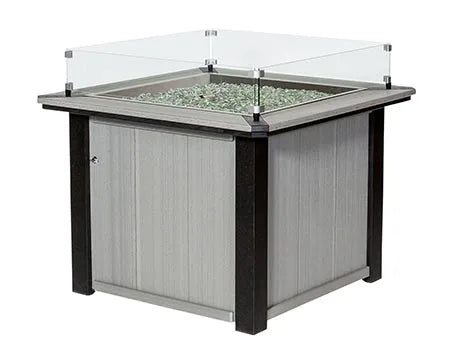  A square fire pit table with a glass top and a glass wind screen. The table is made of gray and black poly lumber and has a flame inside.