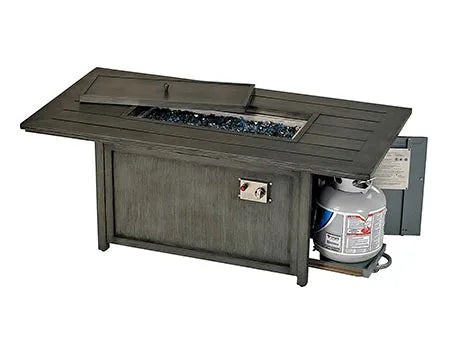 A rectangular outdoor table with fire pit in the middle. It has a gray, wood-like finish. The table has a glass top covering the fire pit, a propane tank storage compartment, and controls for the flames.