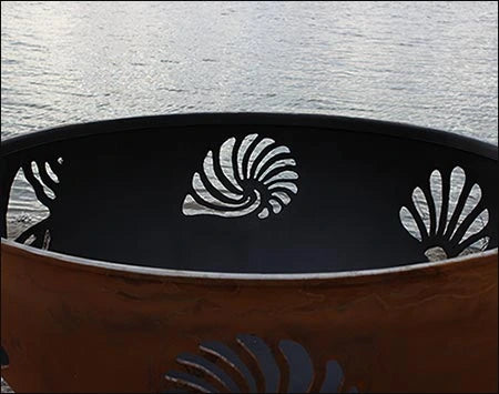  A close-up of a seashell cutout in a fire pit. The seashell is white against a dark background. There is water in the background.