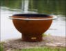 A round, rust-colored wood burning fire pit with a stainless steel handle. It is sitting on a rock near a body of water.