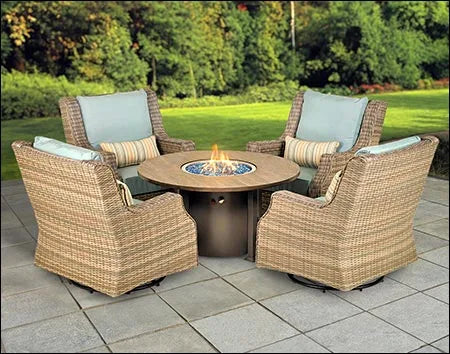 A patio furniture set made of wicker with a circular fire pit table in the center. The set includes four swivel chairs with light blue cushions. The fire pit table has a bronze-colored base and a glass top with blue flames visible. The background shows a green lawn and trees.