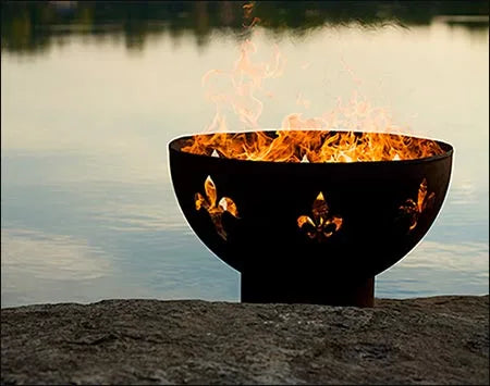 A round, black wood-burning fire pit with fleur-de-lis cutouts. Flames are burning inside the pit, and it is placed on a rock near a body of water.