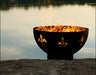 A round, black wood-burning fire pit with fleur-de-lis cutouts. Flames are burning inside the pit, and it is placed on a rock near a body of water.