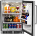 A stainless steel Fire Magic outdoor refrigerator with the door open, revealing the interior. The refrigerator is stocked with various beverages and snacks, including bottles of soda, beer, juice, and cans of soda. There are also fruits like oranges and limes.