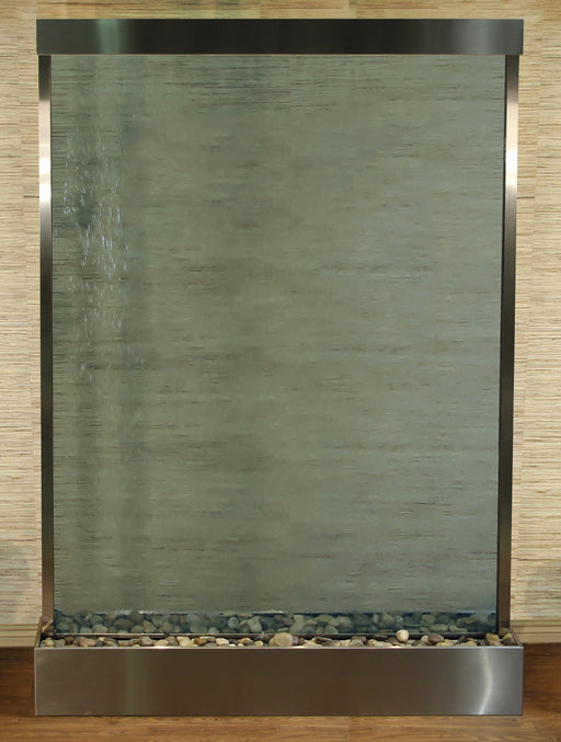 
A towering, rectangular Adagio Mega Grandeur Floor Water Fountain, featuring a clear glass water panel framed by brushed stainless steel. Water flows down the glass, creating a subtly textured, rippling effect across the surface, with a more pronounced cascade visible on the left side. 