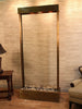 
A tall, rectangular indoor water feature, likely the Adagio Water Features Tranquil River Floor Fountain, stands against a textured light brown wall. The fountain has a dark bronze or copper-toned frame surrounding a wide, vertical panel of what appears to be clear or frosted glass, allowing water to cascade down.
