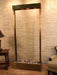 
A tall, rectangular indoor water feature, likely the Adagio Water Features Tranquil River Floor Fountain, stands against a textured light brown wall. The fountain has a dark bronze or copper-toned frame surrounding a wide, vertical panel of what appears to be clear or frosted glass, allowing water to cascade down.
