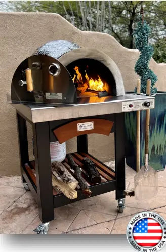 A outdoor pizza oven cart with a black, powder-coated steel exterior and a stainless steel countertop sits on a wheeled cart with a shelf. The oven has a glass window and a chimney. In the background, there is a green lawn and a patio umbrella.