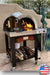 A outdoor pizza oven cart with a black, powder-coated steel exterior and a stainless steel countertop sits on a wheeled cart with a shelf. The oven has a glass window and a chimney. In the background, there is a green lawn and a patio umbrella.