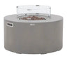 A modern, cylindrical gas fire pit in a solid gray color, featuring a clear, round glass wind guard surrounding the central fire area filled with brown rocks.