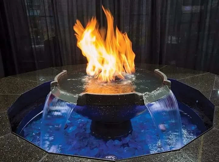 A black, circular fire pit with a water fountain in the center. The fire pit has a metal fire ring and a stainless steel base. The water fountain has a fire burning on top of the water. The fire pit is surrounded by black lava rocks.