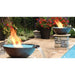 Two fire pits burning next to a swimming pool. The fire pits are circular and made of metal. They are short and have a smooth, reflective surface.