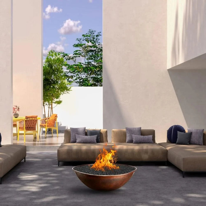 A modern outdoor living spcae with a round metal fire pit in the center. The fire pit is surrounded by a black leather couch and a white armchair. There is a coffee table with a glass top in front of the couch. On the wall behind the couch is a large piece of abstract art.