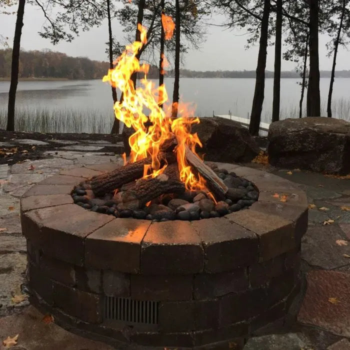 A round, stainless steel fire pit with a lit flame burning in the center. The fire pit is sitting on a slate patio in front of a body of water, possibly a lake. In the background, there are trees and other landscaping.