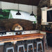 A stainless steel outdoor kitchen countertop with a built-in HPC Villa Series Pizza Oven. The oven has a glass tile front and a stainless steel chimney. There is a wooden table with stools next to the countertop.