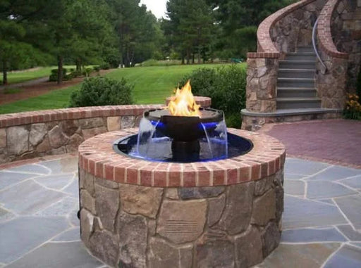 A round, concrete fire pit with a black fire bowl and a stainless steel ring around the top. In the center of the fire pit is a stainless steel gas burner with a large flame burning on the water's surface. The fire pit is surrounded by a shallow, black scupper with four water features flowing down the sides.