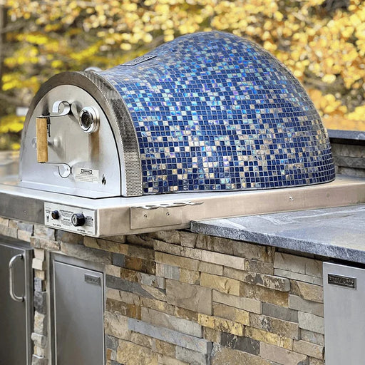 A countertop pizza oven with a blue mosaic tile roof sits on a kitchen counter. The oven has a stainless steel base and a thermometer on the front.