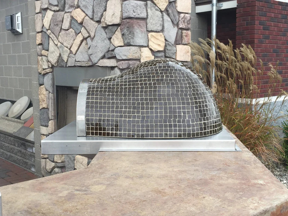 A countertop pizza oven made of stainless steel. It sits on a black countertop next to a fireplace.