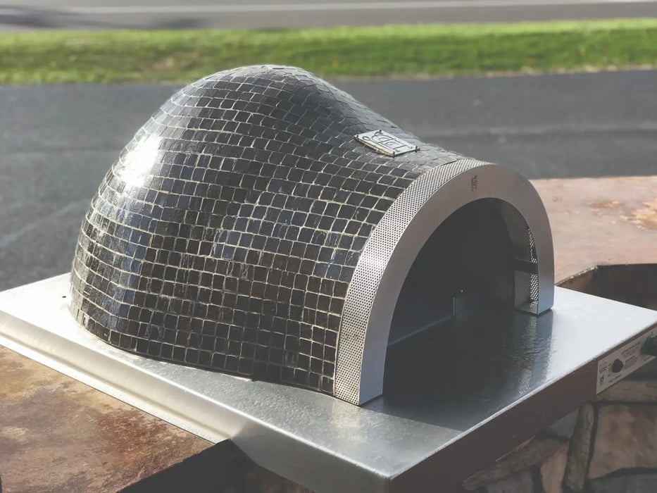 A mosaic tiled pizza oven sitting on top of a stainless steel table. The text on the front of the oven reads "HPC Fire Villa Series Built-In Hybrid Gas/Wood Glass Tile Pizza Oven