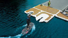 Person on a surfboard near a floating dock with another person standing on a platform.