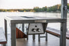 An outdoor grill table with a dark wood top and metal frame, set on a pier or deck overlooking a lake.