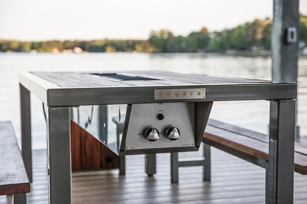 An outdoor grill table with a dark wood top and metal frame, set on a pier or deck overlooking a lake.