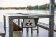 An outdoor grill table with a dark wood top and metal frame, set on a pier or deck overlooking a lake.