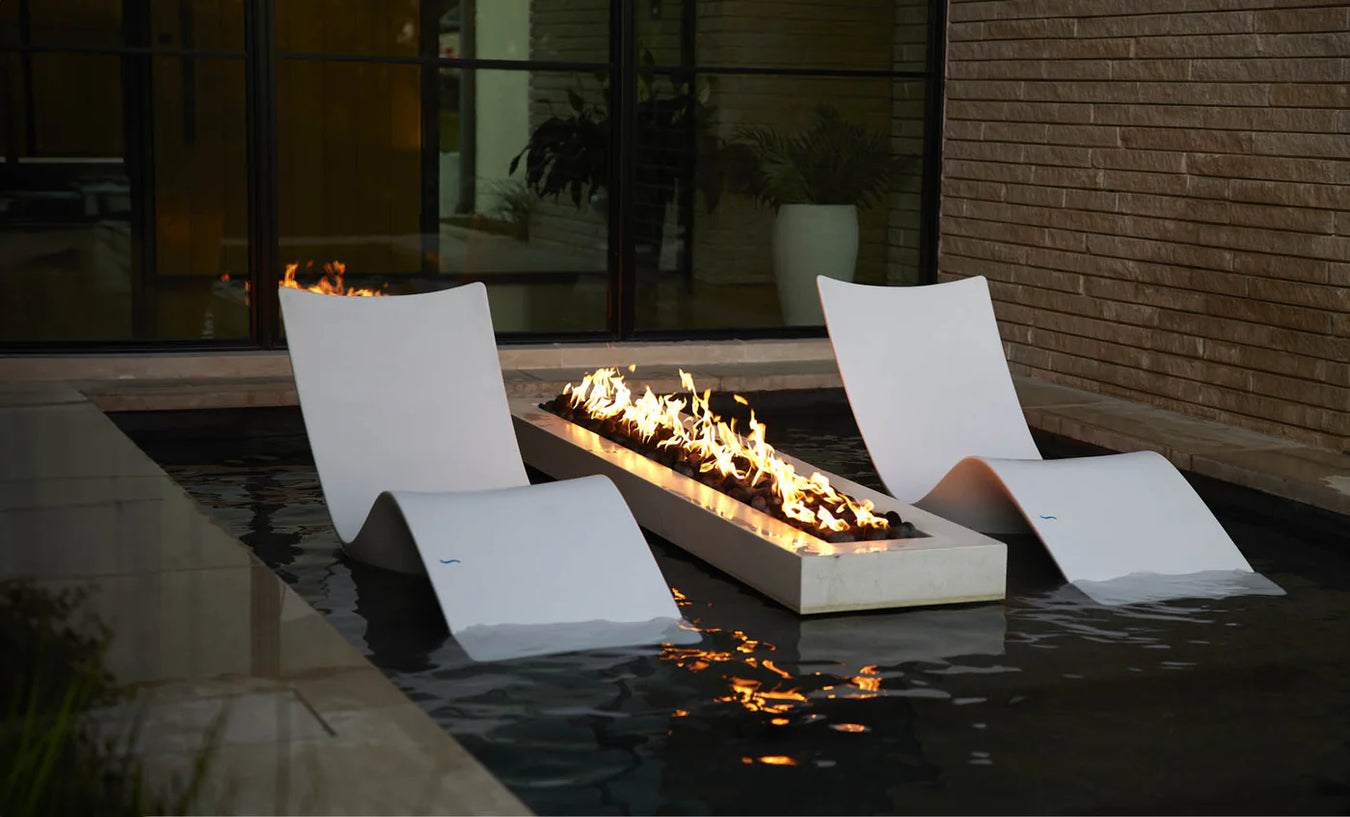 Two white Ledge Lounger chairs partially submerged in shallow water flank a long, rectangular modern fire pit. The fire pit, made of a light-colored material, burns with bright yellow and orange flames and contains dark-colored stones or media.