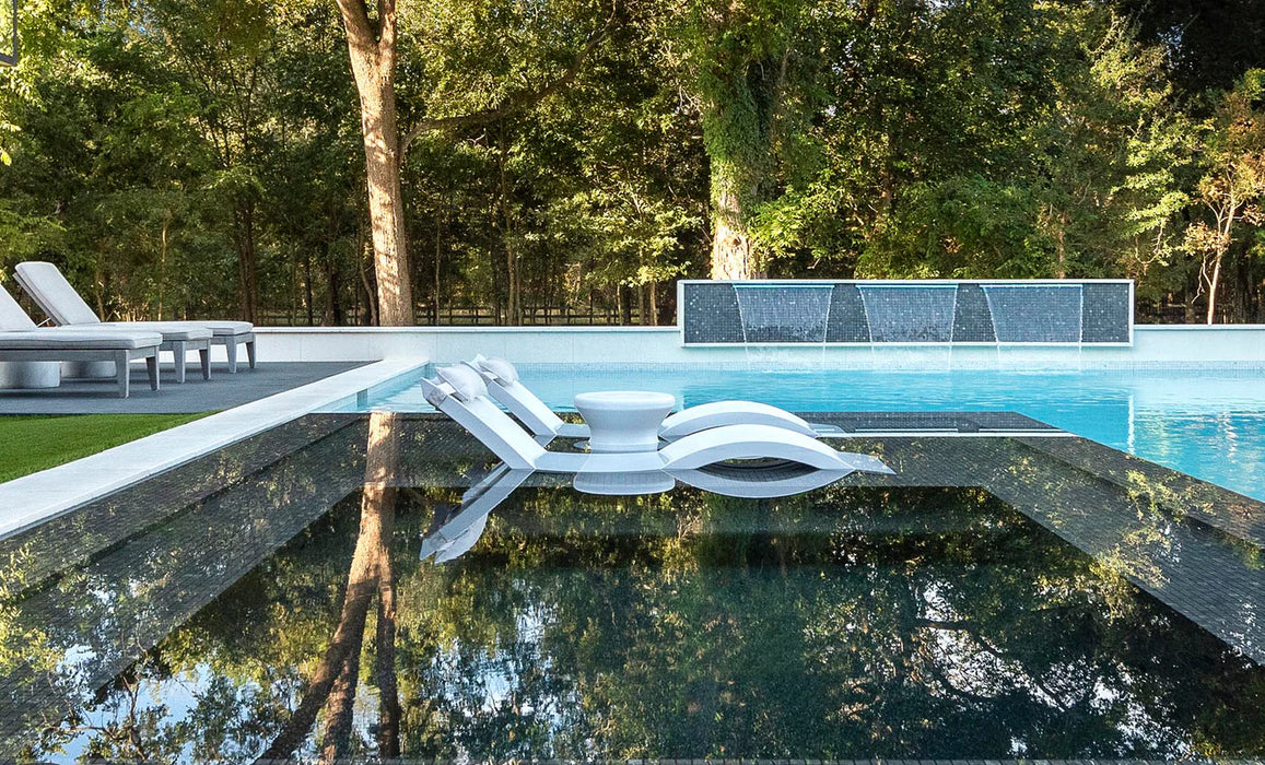 A photo of a luxurious swimming pool with white lounge chairs floating on the water. The pool is surrounded by lush greenery and features a decorative water feature.