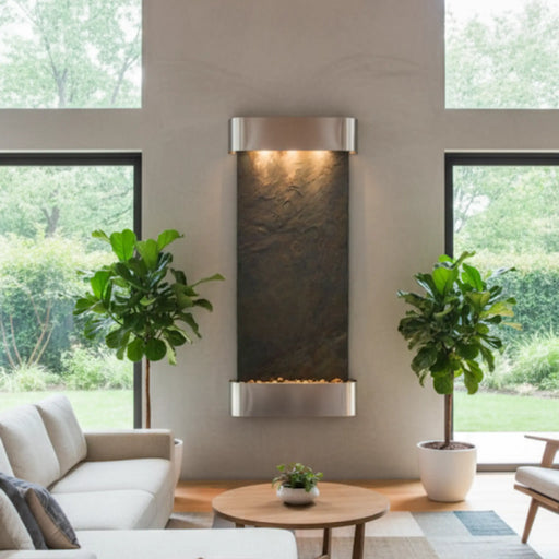 Modern indoor water feature by adagio hung on the wall with plants and a sofa in a room with large windows.