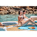Two people relaxing on a sunlit floating yacht platform by a body of water with rocky background