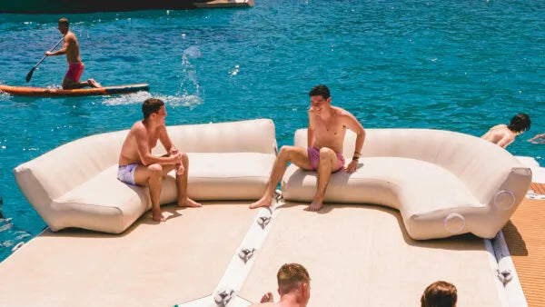People relaxing on inflatable furniture by a body of water with a person paddleboarding.