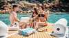 People enjoying a sunny day by the water with drinks and snacks on a wooden deck.