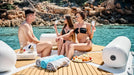 People enjoying a sunny day by the water with drinks and snacks on a wooden deck.