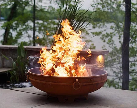 A round, rusted metal fire pit with a large, roaring fire burning inside. The fire pit has two handles on the side and is placed on a stone patio. There is a body of water and lush greenery in the background.